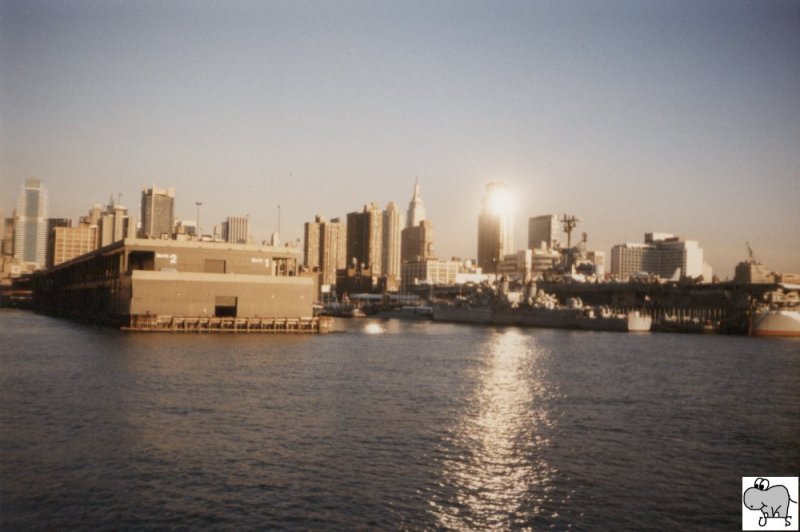Die tiefstehende Sonne spiegelt sich in einen Wolkenkratzer auf Manhatten. Im Vordergrund ist das  Intrepid Sea Air Space Museum  zu sehen. Die Aufnahme stammt von 1997.