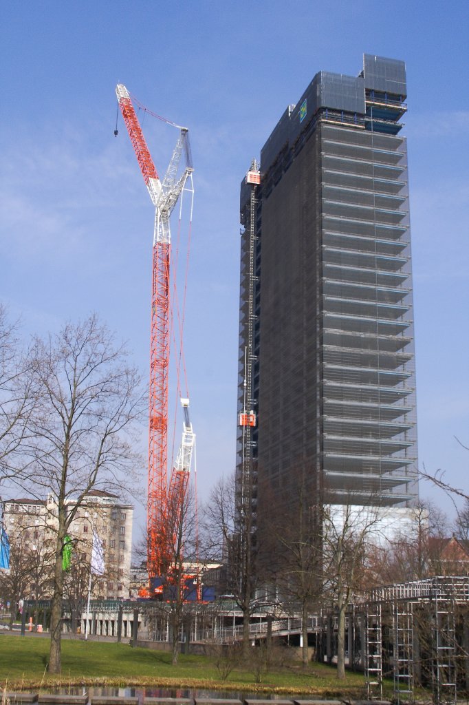 Der riesige Baukran  zerpflckt  nach und nach das im Rckbau befindliche Bayer-Hochhaus in Leverkusen! Gut ist hier bereits das Fehlen der ersten Segmente im oberen Gebudebereich zu erkennen. Die Aufnahme erfolgte am 24.03.2012 vom im Carl-Duisberg-Park gelegenen BayKomm (Bayer-Kommunikationszentrum) aus.