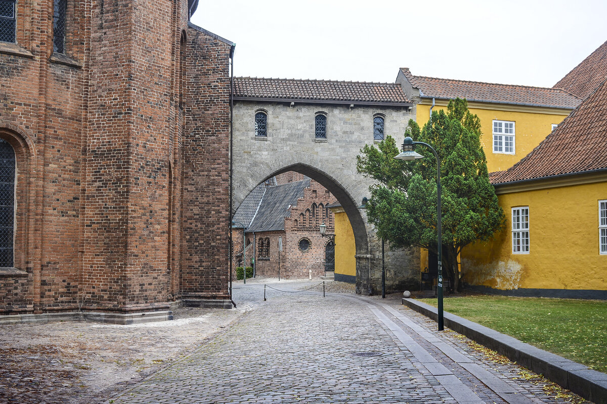 Absalonsbuen am Domplatz von Roskilde. Das Backsteingebude auf der linken Seite ist Teil des Domvon Roskilde. Das gelbe Gebude auf der rechten Seite ist Teil des Museums fr zeitgenssische Kunst(dnisch: Museet for Samtidskunst)  in Roskilde.
Aufnahme: 20. Juni 2023.