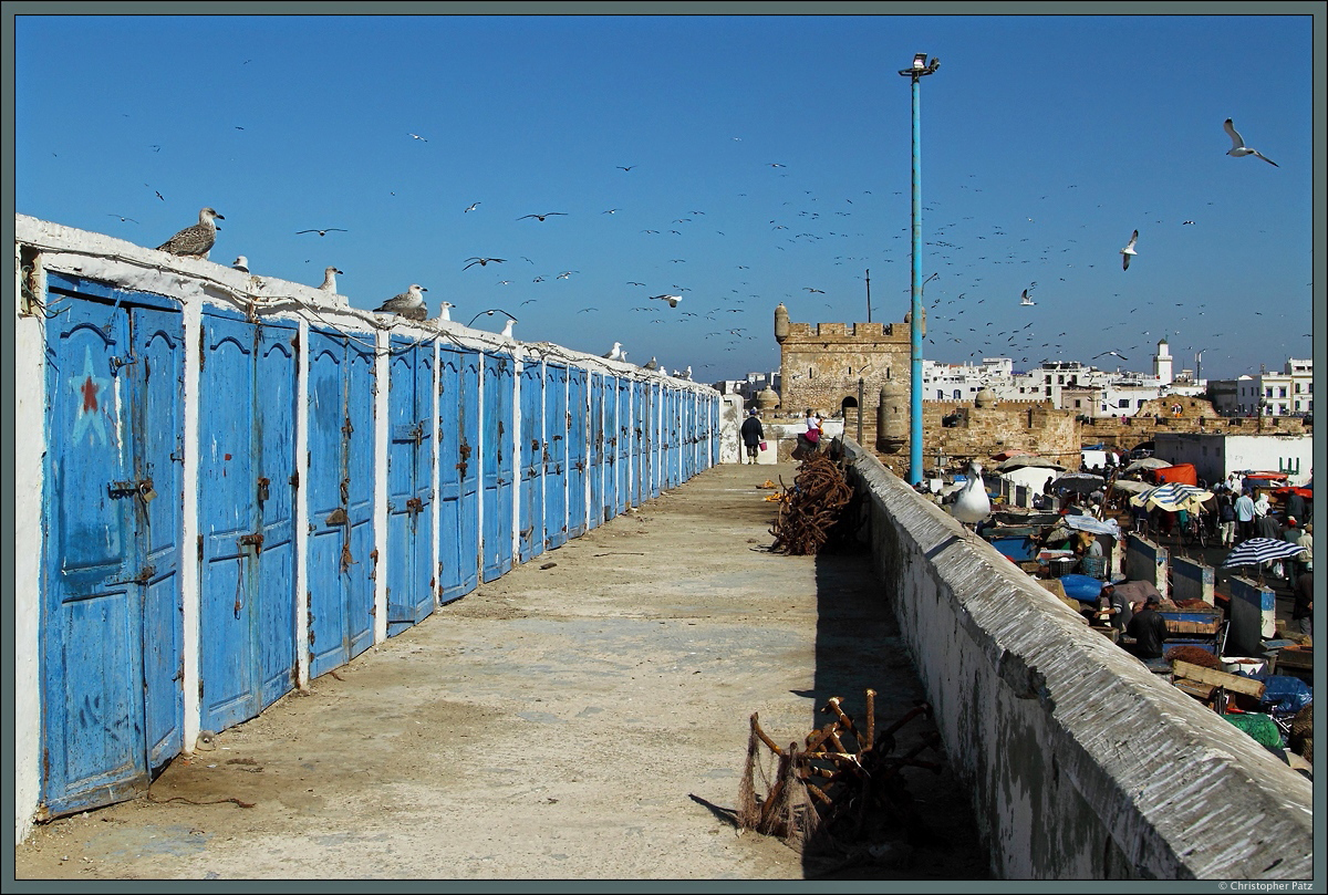 Auf der alten Hafenmauer von Essaouira befinden sich die Lager der Fischer, die im Hafen ihren Fang verkaufen. Im Hintergrund sind die Festungsanlage Scala du Port und die Medina von Essaouira zu sehen. (21.11.2015)