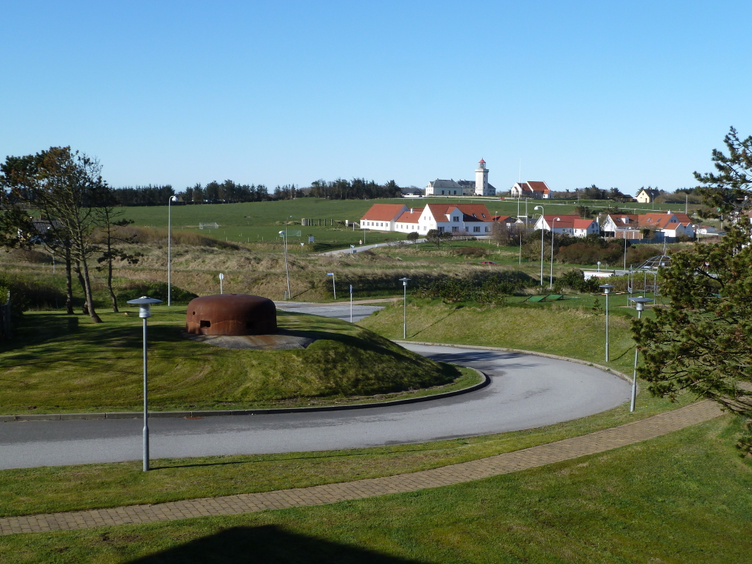 Blick am 18.04.2014 ber den kleinen Ort Hanstholm zum Leuchturm. Im Vordergrund ist ein kleiner Bunker zu sehen, von denen es in Hanstholm und Umgebung sehr viele gibt, da die Stadt und ihr Umfeld zu Zeiten des II. Weltkrieges von der damaligen Deutschen Wehrmacht als riesige Festungsanlage ausgebaut wurde, welche dem Schutz der hier befindlichen II. Kstenbatterie diente. Zu diesem Zweck wurden smtliche Einwohner zwangsevakuiert, die Gegend weitrumig abgesperrt und strengstens bewacht. Heute ist Hanstholm ein ruhiger, idyllischer Kstenort, mit Fischerei- und Fhrhafen, sowie einer Fischfabrik. Sehr sehenswert ist das im Ort befindliche Museumscenter, mit Bunkermuseum, Kanonenausstellung, riesigen Geschtzstnden, sowie den berresten der ehemaligen Munitionsbahn, auf deren Gleisen heute ein kleiner Besichtigungszug verkehrt, mit welchem man sogar durch Munitions- und Geschtzbunker fahren kann. (Bildautor: Jrg-Uwe Bhle, Genehmigung zur Verffentlichung liegt vor!)