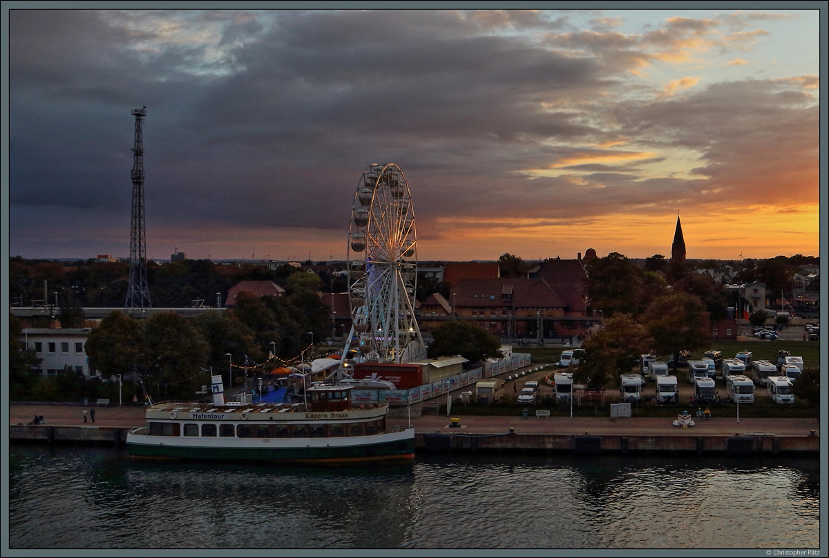 Blick vom Schiff auf Warnemnde: Zwischen Warnow und Bahnhof Warnemnde leuchtet ein Riesenrad im Abendlicht. Davor liegt die  Kpp'n Brass  am Anlieger. (25.09.2021)