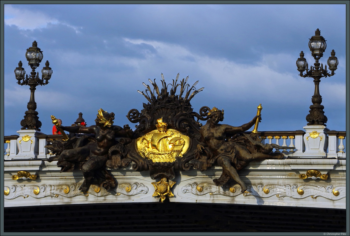 Die Pont Alexandre III, eine 1897 errichtete neobarocke Brcke ber die Seine, ist mit zahlreichen eindrucksvollen Figuren geschmckt. Hier ist das von zwei Seine-Nymphen flankierte Pariser Stadtwappen zu sehen. (17.07.2018)