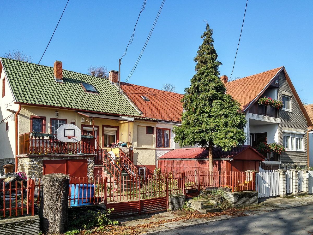 Dieses Foto habe ich in Pcs, in der Stadtteil  Erzsbettelep  (Elisabethsiedlung) gemacht. Dieser Stadteil wurde durch die Donaudampfschiffgesellschaft gegrndet. Die  Kolonie  stand aus 4 Strassen und 32 Huser. Es gab 6 kleine Wohnungen pro Haus zzgl Gartenteil und Nebengebude. Vor ca. 100-120 Jahren wurden neue Kohlenminien in der Nhe erffnet und die dort arbeitenden haben hier Wohnungen bekommen. Die Siedlung hie ursprnglich Ullmannsiedlung, genannt nach einem Leiter des Unternehmens. 1938 wurde auf Elisabethsiedlung umbenannt (von Elisabeth Amalie Eugenie von Wittelsbach Knigin genannt). Die Huser waren berdurchschnittlich ausgerstet, da Leitungswasser und Strom auch vorhanden war. In den vergangenen guten Jahrhundert wurde es nicht geregelt wie man die Gebuden/Wohnungen ndern, erweitern, umbauen oder zusammenlegen/vereinigen darf. Deswegen ist auf heutzutage das Strassenbild ganz ganz gemischt (und daher meiner Meinung nach, sehr einzigartig und interessant) geworden. Die Etagen wurden gendert, Dachgeschosse wurden eingebaut...daher findet man hier keine zwei gleiche Huser. Die groe nderungen sind um ca 1926 begonnen. Wegen der damaligen Wohnungsmangel hat die Stadt 2/3-tel der Siedlung in Miete genommen und hat unter anderem eine Kirche und Schule erbaut und geffnet. Aufnahmezeit: November 2020.
Garage und Frontpertei sind hchstwahrscheinlich Nachbau. Diese immobilien scheinen richtig Instand gehalten zu sein, und regelmaig / neulich saniert zu sein.