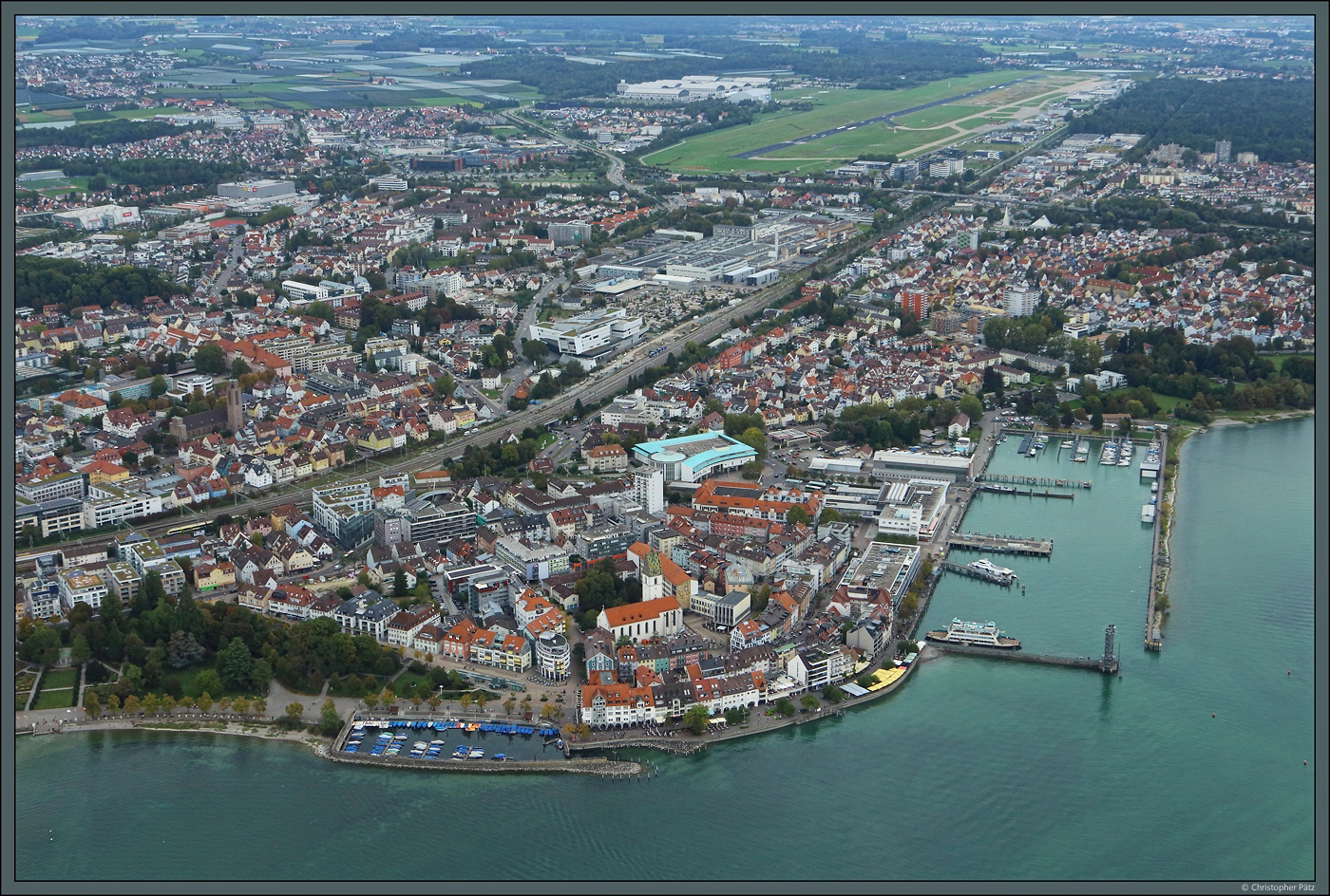 Am Seeufer liegt die Altstadt von Friedrichshafen, mit der Kirche St. Nikolaus im Zentrum. Links der Bahngleise ist die Kirche St. Petrus-Canisius zu erkennen. Im Hintergrund ist der Flughafen zu sehen, zwischen Stadt und Flughafen wiederum die Hallen des ZF-Werkes Friedrichshafen. (05.10.2023)