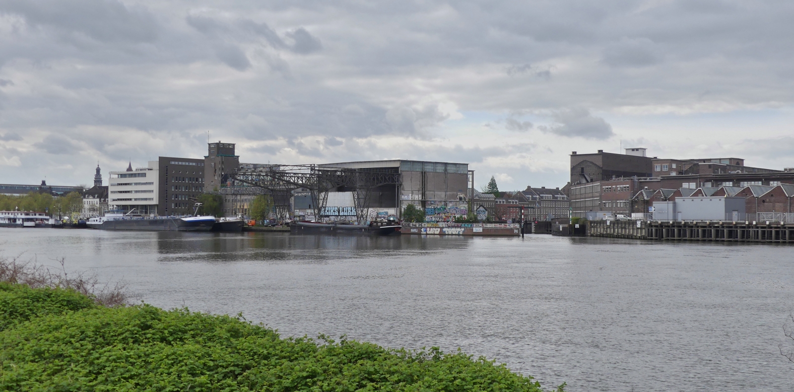 Blick von der Rechten Maas Seite auf die frheren Lagerhallen fr die Landwirtschaft, heute ein Aufenthaltsort fr Knstler und sonstige Vereine, am rechten Bildseite ist die Schleuseneinfahrt zum Binnenhafen in Maastricht zusehen. 04.2024