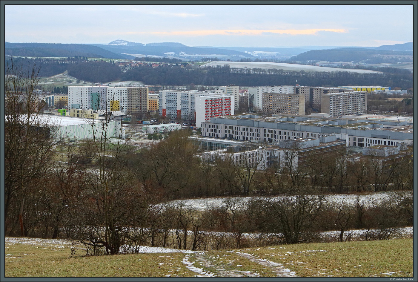 Der Stadtteil Jena-Lobeda entstand zwischen 1966 und 1988 westlich des lndlich geprgten Stadtteils Lobeda-Altstadt als Plattenbausiedlung. In der rechten Bildhlfte sind die Gebude des Universittsklinikums zu sehen, dahinter die Hochhuser von Lobeda-Ost. Auf dem gegenberliegenden Hgel liegt das Dorf Sulza, dahinter ist auf einem Berggipfel am Horizont die Leuchtenburg zu erahnen. (Jena, 14.01.2024)