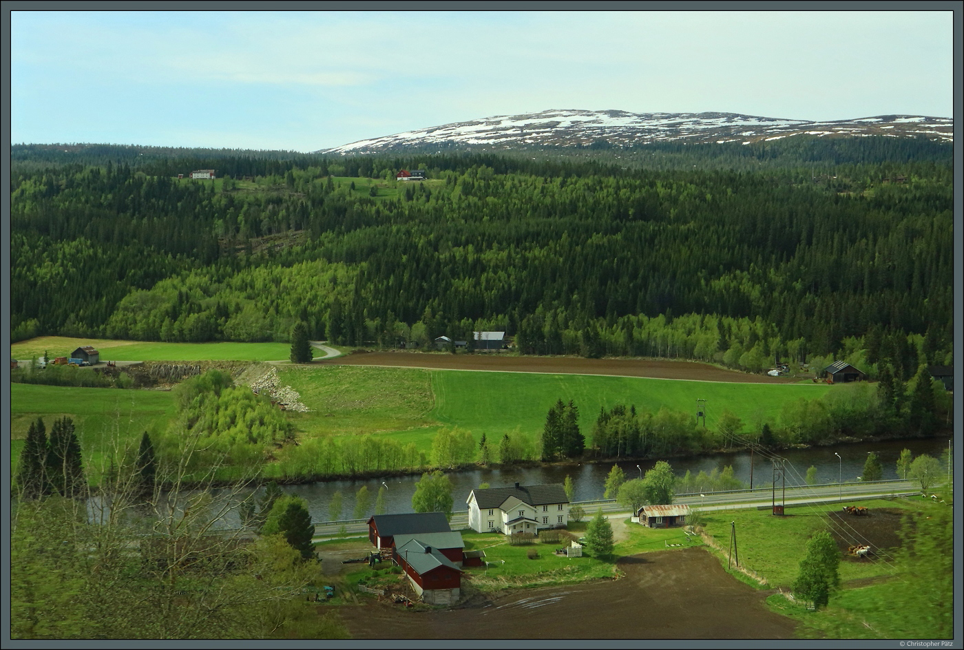 Entlang dem Fluss Gaula reihen sich mehrere Gehöfte des Ortes Haltdalen. Im Hintergrund ist der 956 m hohe Berg Gråmerra zu sehen, der auch am 27.05.2023 hoch teilweise schneebedeckt ist.