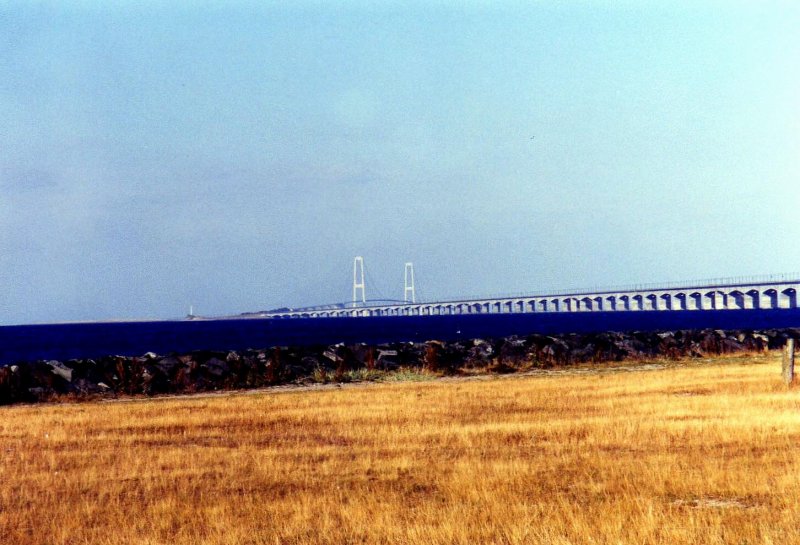Hierbei handelt es sich um die Storebeltbrücke zwischen der Insel Fünen und der Insel Seeland. Der Auftrag zum Bau der Brücke über den Großen Belt(Storebelt) wurde am 12. Juni 1986 vom dänischen Parlament erteilt. Am 14 Juni 1998 wurde das Bauwerk für den Verkehr freigegeben. Von der Insel Fünen aus, bei der Stadt Nyborg, führt zuerst eine knapp 7 Km lange Auto-und Eisenbahnbrücke auf die kleine Insel Sprogø. Die Straße besitzt jeweils 2 Fahrspuren in jede Richtung und die Bahntrasse verläuft zweigleisig. Von der Insel Sprogø aus überquert die Autobahn mit einer weiteren knapp 7 Km langen Brücke die Ostsee bis zur Insel Seeland, nahe der Stadt Korsør, während die Bahntrasse ab der Insel Sprogø in einem 8 Km langen Bahntunnel bis zur Insel Seeland verschwindet. Die beiden Pylonen des Brückenteils von Sprogø nach Seeland bilden mit knapp 250m den höchsten Punkt Dänemarks. Bei sehr extremen Wetterbedingungen kann es passieren das die Brücke für den Verkehr zeitweise gesperrt werden muss. Das Bild enstand bei Nyborg auf Fünen (gescanntes Bild)