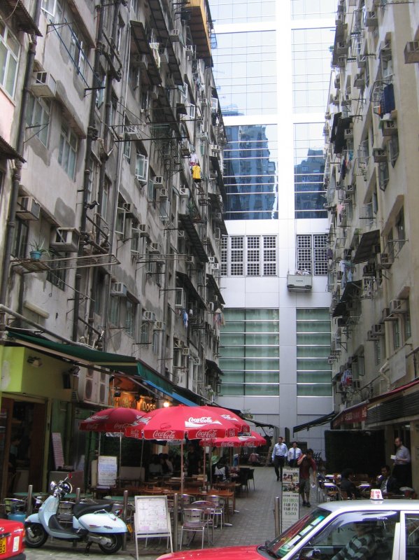 Hong Kong : Eine Idylle der Tristesse und der Gegenstze. Im Hintergrund ein modernes Geschftsgebude, davor die schbigen Mietskasernen und unten in den Durchgang drngt sich ein kleines Restaurant.  Nur nicht nach oben gucken , mchte man den beiden Geschftsleuten zurufen die das Restaurant besuchen mchten.