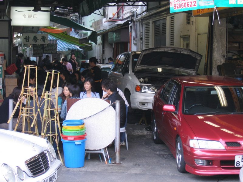 Hong Kong : Raum ist in der kleinsten Htte! Die jungen Leute sitzen an den Tischen eines kleinen Restaurants in direkter Nhe neben einer Autowerkstatt. Dieses Idyll fand ich in einer Seitenstrasse in der Nhe des Somerset House in Quarry Bay.