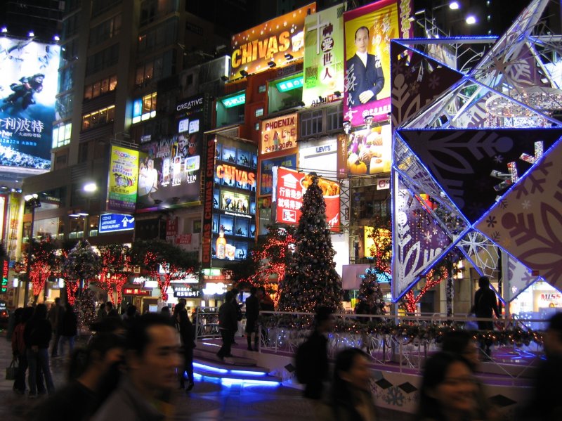 Hong Kong bei Nacht. Der Lichterglanz des Weihnachtsbaums vor dem Times Square Center wirkt sehr sparsam gegen die grell-helle Lichtreklame im Scheinwerferlicht.