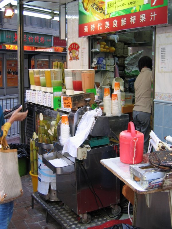 Hong Kong - Szenen auf einem Strassenmarkt zwischen Hopewell Center und Hennessy Rd. 2,5 qm reichen aus, um bereits einen Saftladen zu betreiben. Die Dame hat schon das Portemonaie gezckt. Welchen der bunten Sfte wird sie wohl whlen? Und was ist wohl in der Gieskanne?