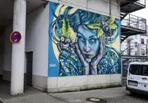 Stine Hvid ist eine d�nische K�nstlerin, die schon fr�h ihre ganz eigene Bildsprache entwickelt und f�r ihre Kunst gefunden hat. Bei ihrem Motiv f�r die Nord:Syd Freiraumgalerie in der Flensburger Nikolaistra�e handelt es sich um ein zauberhaftes Portr�t einer jungen Frau, umgeben von zwei magischen Wesen, die sie umsorgen. Der Stil des Bildes l�sst sich als magischer Realismus bezeichnen. Aufnahme: 26. Juli 2026.