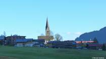 Die katholische Pfarrkirche St. Jodok in Mittelberg, Kleinwalsertal, und das Dach eines der Huser in der Nhe werden bereits von der Sonne angeleuchtet; die restlichen Huser liegen am Morgen noch im Schatten (Bezirk Bregenz, Land Voralberg/sterreich, Oktober 2013).