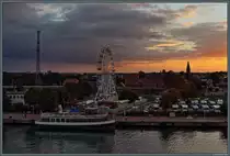 Blick vom Schiff auf Warnem�nde: Zwischen Warnow und Bahnhof Warnem�nde leuchtet ein Riesenrad im Abendlicht. Davor liegt die  K�pp'n Brass  am Anlieger. (25.09.2021)